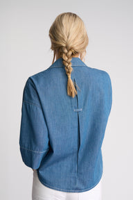 A woman viewed from the back wearing a blue denim shirt jacket with three-quarter sleeves and a central seam detail, paired with white pants against a light gray studio background.