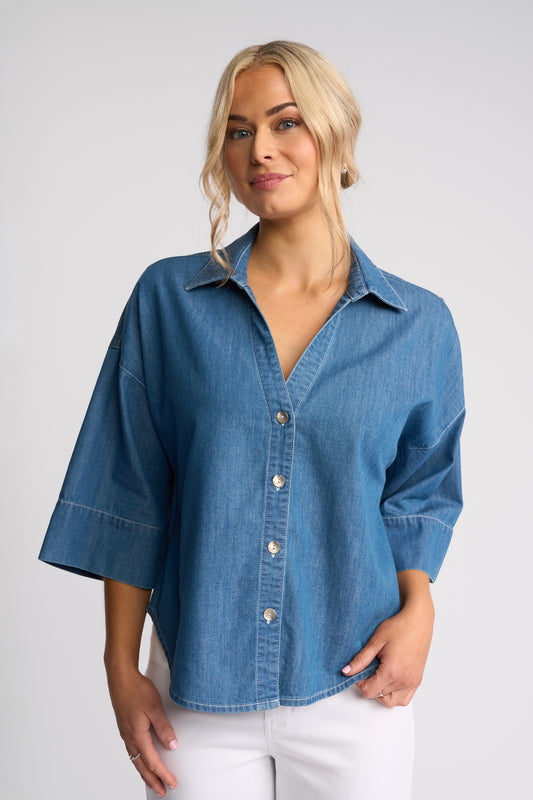 A woman wearing a blue denim button-up shirt with three-quarter sleeves and white pants, photographed against a light gray studio background.