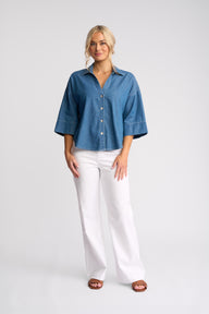 A woman modeling a blue short-sleeve button-up shirt with white wide-leg pants and brown sandals, standing against a light gray studio background.