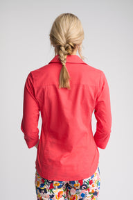 A woman viewed from behind wearing a coral red long-sleeve button-up shirt and colorful floral pants against a light gray studio background.