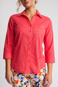 A woman wearing a coral pink button-up shirt with three-quarter sleeves and two chest pockets, paired with colorful floral pants, against a light gray studio background.