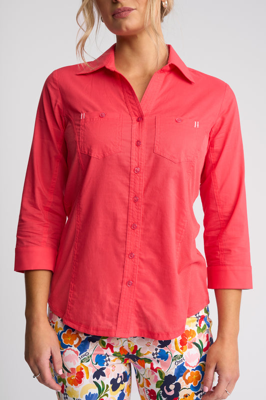 A woman wearing a coral pink button-up shirt with three-quarter sleeves and two chest pockets, paired with colorful floral pants, against a light gray studio background.