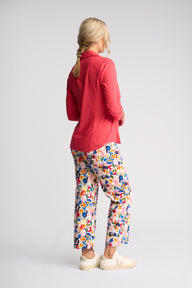 A woman shown from the back wearing a red long-sleeve button-up shirt and colorful abstract-print pants, paired with cream slip-on shoes, against a light gray studio background.