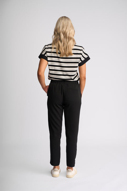 A woman viewed from behind wearing a black and white horizontally striped short-sleeve top, black trousers, and white sneakers, standing against a light gray studio background.