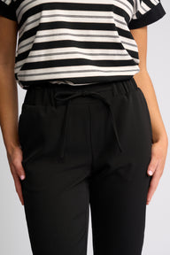 A close-up of a person wearing black drawstring trousers and a black-and-white striped top against a light gray studio background.