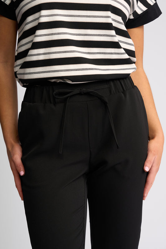A close-up of a person wearing black drawstring trousers and a black-and-white striped top against a light gray studio background.