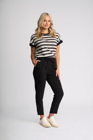 A woman modeling a black and white horizontally striped short-sleeve t-shirt, paired with black cropped trousers and white sneakers, against a light gray studio background.