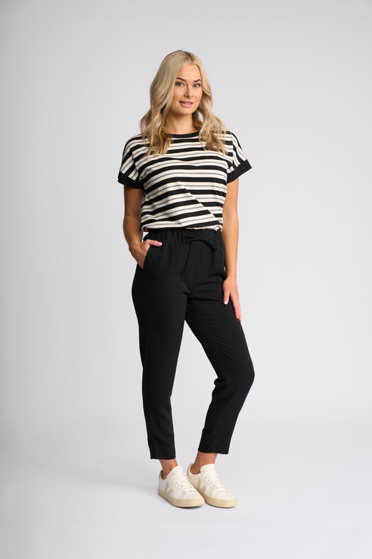 A woman modeling a black and white horizontally striped short-sleeve t-shirt, paired with black cropped trousers and white sneakers, against a light gray studio background.