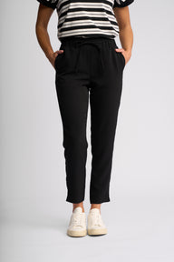 A person wearing black drawstring ankle-length pants with side pockets, paired with a black-and-white striped top and white sneakers, standing against a light gray studio background.