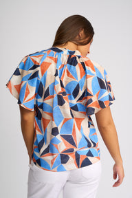 A woman viewed from the back wearing a short-sleeve blouse with a bold geometric pattern in blue, orange, beige, and navy, paired with white pants, against a light gray studio background.