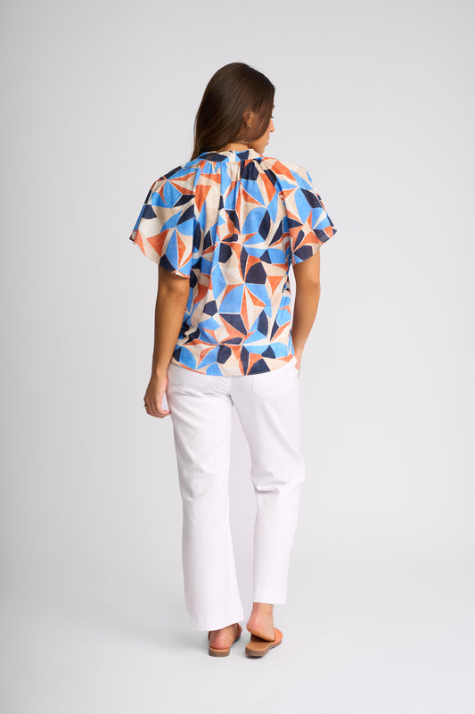A woman viewed from the back wearing a short-sleeve blouse with a colorful geometric pattern in blue, orange, beige, and black, paired with white pants and brown sandals, against a light gray studio background.