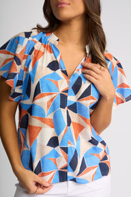 A woman wearing a short-sleeve button-front blouse with a bold geometric pattern in blue, orange, and navy on an off-white background, paired with white pants against a light gray studio backdrop.