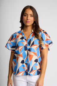 A woman modeling a short-sleeve button-up blouse with a vibrant geometric pattern in blue, orange, and cream, paired with white pants against a neutral gray studio background.