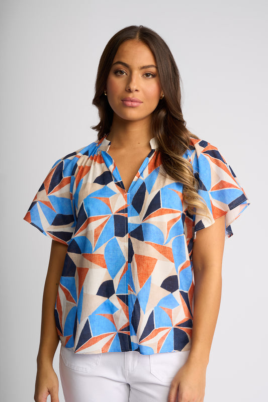 A woman modeling a short-sleeve button-up blouse with a vibrant geometric pattern in blue, orange, and cream, paired with white pants against a neutral gray studio background.