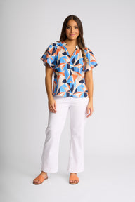 A woman modeling a short-sleeve button-up blouse with a vibrant geometric pattern in blue, orange, and black, paired with white pants and brown sandals, against a light gray studio background.