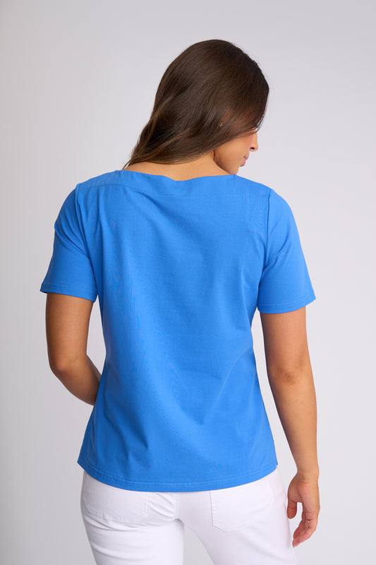 A woman viewed from the back wearing a bright blue short-sleeve crewneck t-shirt and white pants, set against a plain light gray studio background.