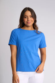A woman modeling a bright blue short-sleeve top with a subtle wavy texture, paired with white pants, against a light gray studio background.
