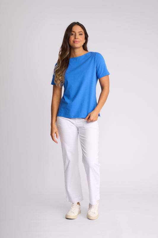 A woman modeling a bright blue short-sleeve crewneck t-shirt, paired with white pants and white sneakers, standing against a light gray studio background.