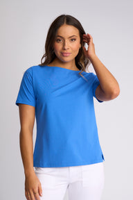 A woman modeling a bright blue short-sleeve boat-neck top with white pants, standing against a light gray studio background.