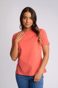A woman modeling a coral short-sleeve crewneck t-shirt with blue jeans, standing against a light gray studio background.