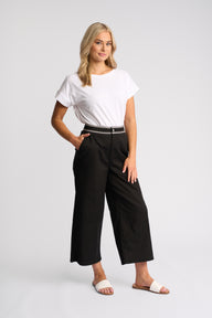 A woman modeling black wide-leg cropped pants with a thin black belt featuring a silver-tone chain detail, paired with a white t-shirt and white slide sandals, against a light gray studio background.
