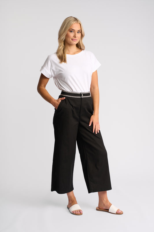 A woman modeling black wide-leg cropped pants with a thin black belt featuring a silver-tone chain detail, paired with a white t-shirt and white slide sandals, against a light gray studio background.