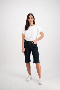 A female model wearing Vassalli indigo denim shorts with contrast stitching, a white cropped top, and white sneakers.