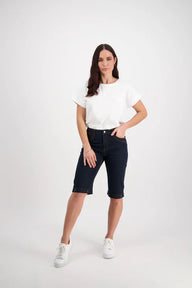 A female model wearing Vassalli indigo denim shorts with contrast stitching, a white cropped top, and white sneakers.