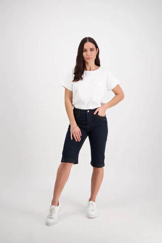A female model wearing Vassalli indigo denim shorts with contrast stitching, a white cropped top, and white sneakers.