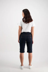 A female model wearing Vassalli indigo denim shorts with contrast stitching, a white cropped top, and white sneakers.