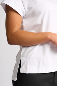 Close-up of a person wearing a white short-sleeve scrub top with a side slit and pocket detail, paired with black bottoms, against a light gray background.