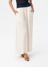 A model wearing a pair of off-white, wide-leg maxi pants with side pockets, paired with a dark top and brown flat sandals, shown from the waist down against a plain white background.