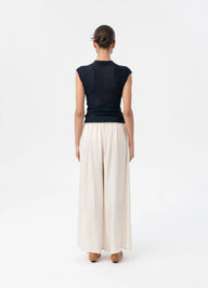 A woman viewed from the back wearing a dark navy sleeveless knit top and cream wide-leg pleated trousers, standing against a plain white studio background.