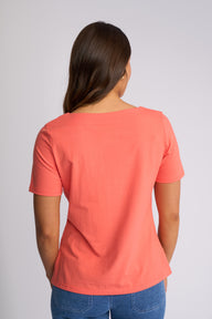 A woman viewed from the back wearing a coral orange short-sleeve t-shirt and blue jeans, standing against a light gray studio background.