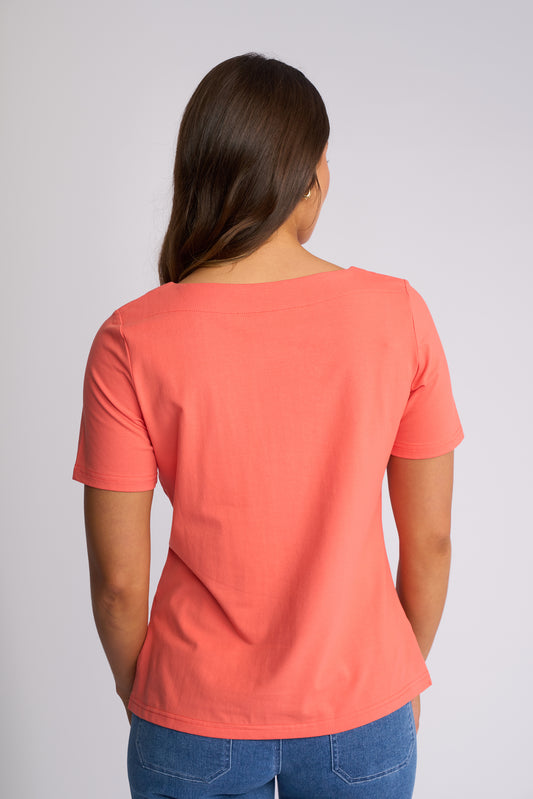 A woman viewed from the back wearing a coral orange short-sleeve t-shirt and blue jeans, standing against a light gray studio background.