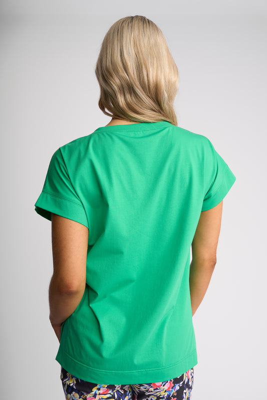 A woman viewed from behind wearing a bright green short-sleeve t-shirt and patterned pants against a light gray studio background.