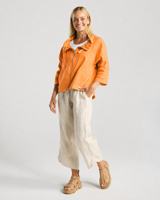 A woman modeling a bright orange zip-up jacket over a white top, paired with light beige linen wide-leg pants and tan platform sandals, against a light gray studio background.
