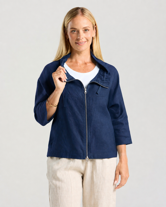 A woman modeling a navy zip-up jacket over a white top, paired with light beige linen wide-leg pants and tan platform sandals, against a light gray studio background.