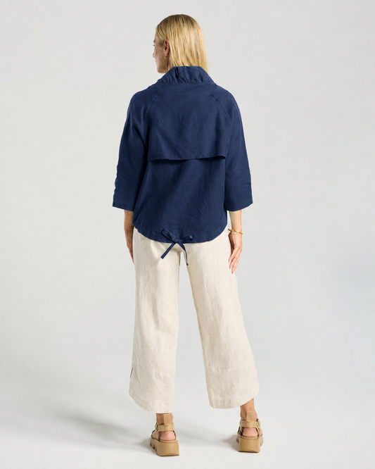 A woman modeling a navy zip-up jacket over a white top, paired with light beige linen wide-leg pants and tan platform sandals, against a light gray studio background.