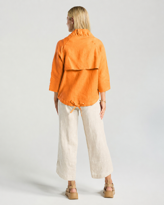 A woman modeling a bright orange zip-up jacket over a white top, paired with light beige linen wide-leg pants and tan platform sandals, against a light gray studio background.
