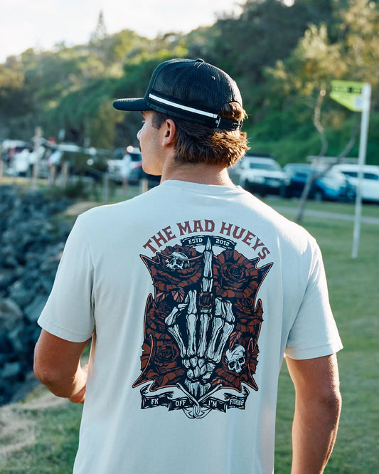 A man viewed from behind wearing a white t-shirt with a large red and black graphic print on the back that reads 'THE MAD HUEYS' and '1979 - 2025'. He is also wearing a black baseball cap with a white stripe.