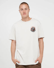 A man wearing a cream-colored short-sleeve t-shirt with a circular graphic on the left chest, paired with brown pants, against a plain light gray background.