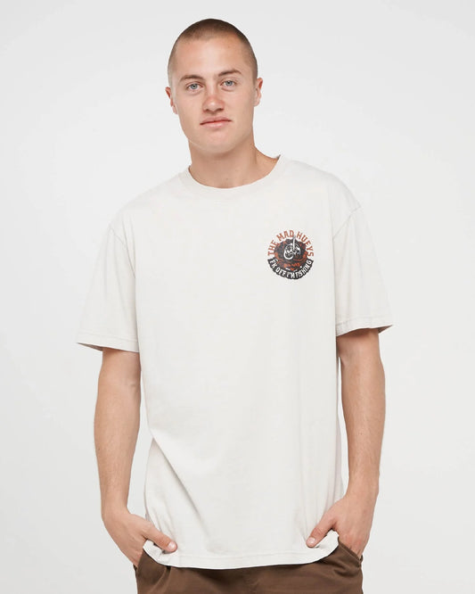 A man wearing a cream-colored short-sleeve t-shirt with a circular graphic on the left chest, paired with brown pants, against a plain light gray background.