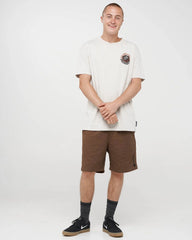 A man modeling a white crewneck t-shirt with a small circular graphic on the left chest, paired with brown shorts, dark gray socks, and black skate shoes, against a plain white studio background.
