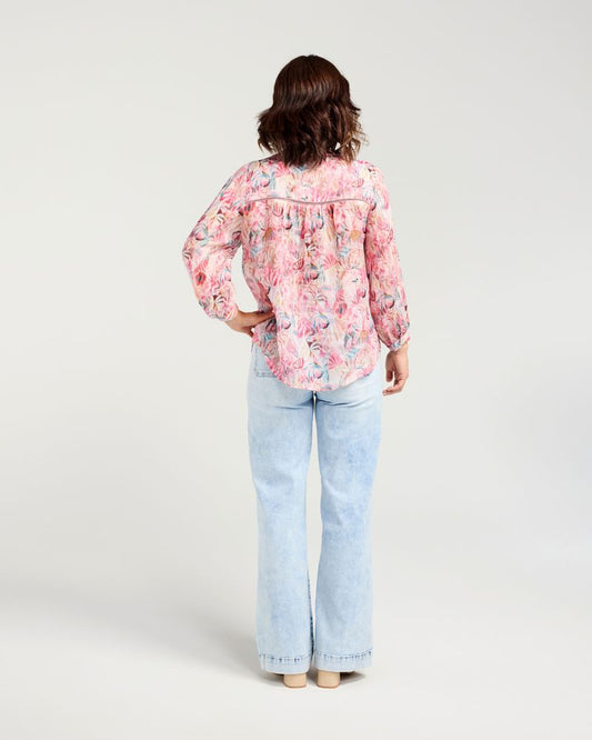 A woman shown from the back wearing a pink floral long-sleeve blouse and light blue jeans, standing against a plain light gray studio background.