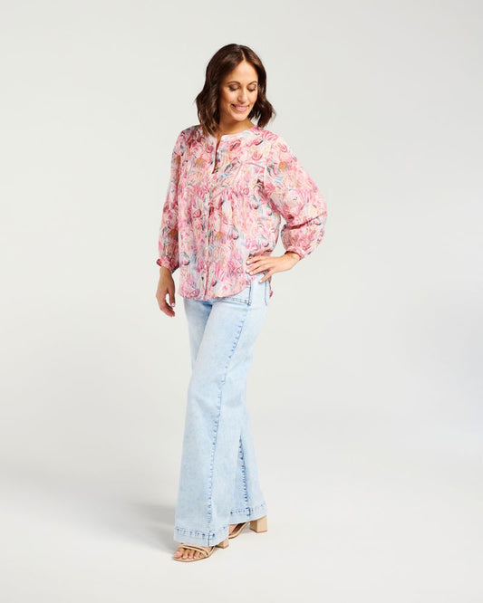 A woman modeling a pink floral long-sleeve blouse with a gathered waist, paired with light-wash flared jeans and beige sandals, against a plain light gray studio background.