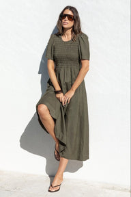 A woman wearing an olive green midi dress with puff sleeves and a smocked bodice, posing against a white textured wall.
