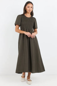 A woman modeling a dark olive green midi dress with short puff sleeves, a smocked bodice, and a flared skirt, paired with white platform shoes against a plain white studio background.