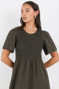 A woman wearing an olive green short-sleeve dress with a smocked bodice and puffed sleeves, photographed against a plain light background.