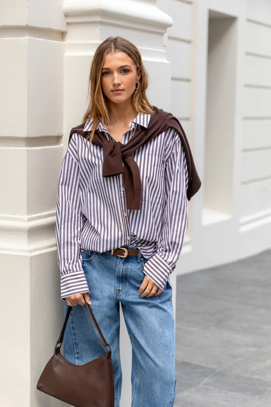 Woman wearing a striped shirt with a brown scarf and blue jeans, standing against a light-colored wall.